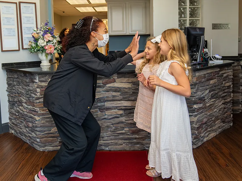 Dental office staff member wearing a face mask giving a high five to two young girls in a welcoming reception area.